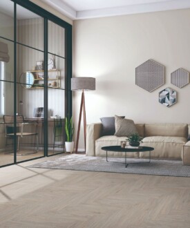 Salón moderno con suelo en espiga SPC Herringbone Taupe de TRIP The Natural Company, sofá beige, mesa de centro redonda, lámpara de pie y zona de trabajo tras cristalera negra.